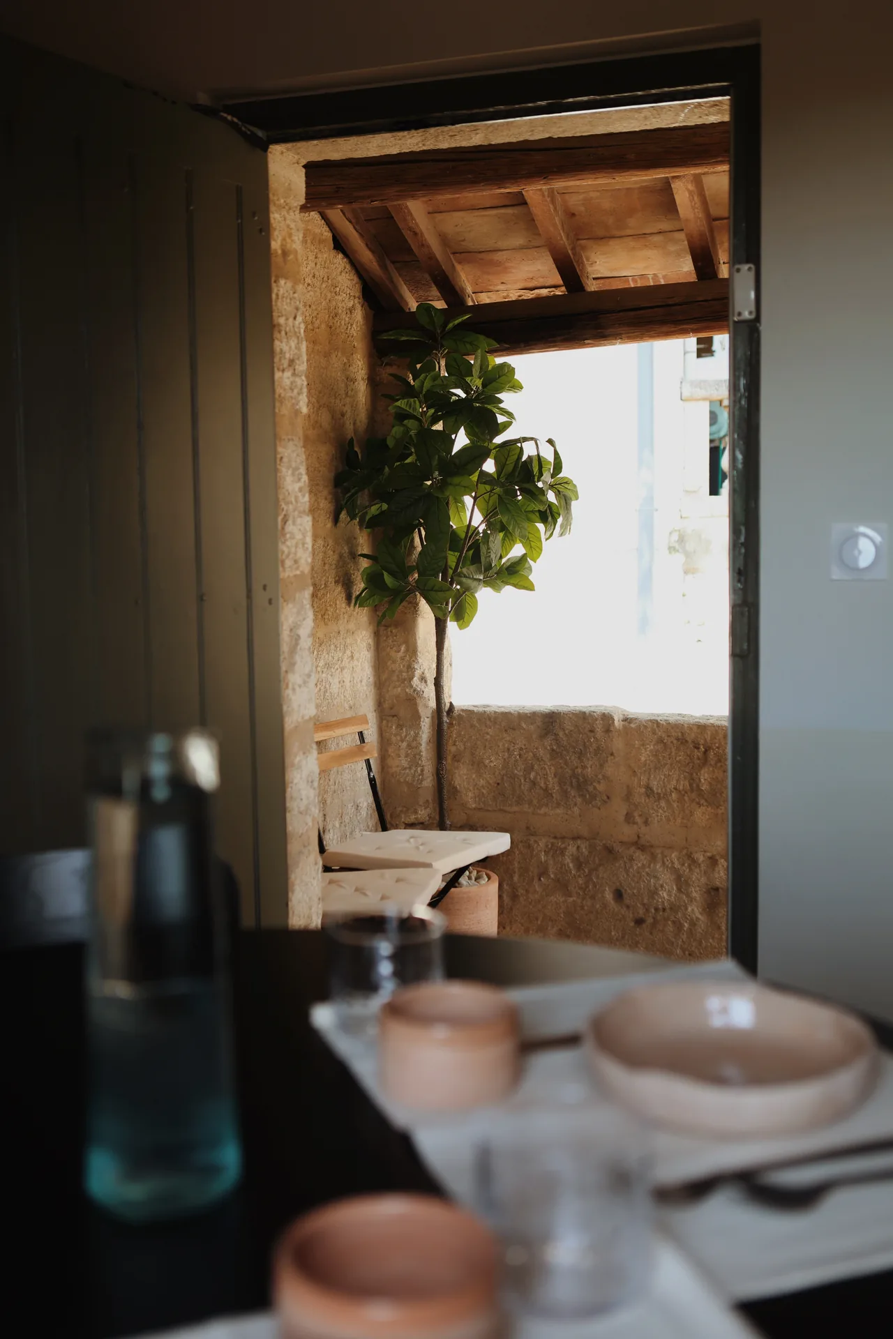 Balcony seen through a dark green door, with a potted tree, a folding chair and ceramic tableware