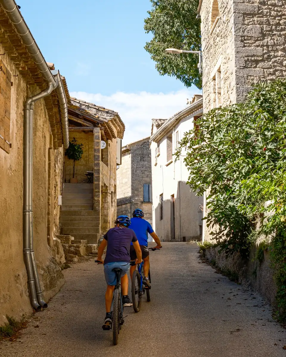 Two cyclists riding up a stone lane in the village, with a fig tree and sunlit limestone houses