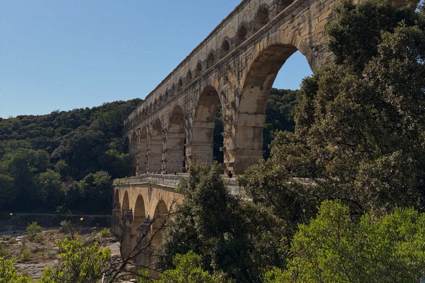 The stone arches of the Pont du Gard aqueduct rising above green trees on the river bank, under a clear blue sky