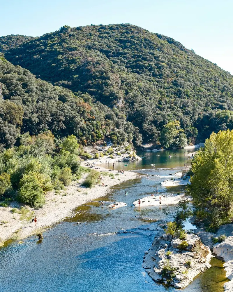 A river in the Gard with pale gravel banks and white rock slabs below wooded hills, with swimmers in the water