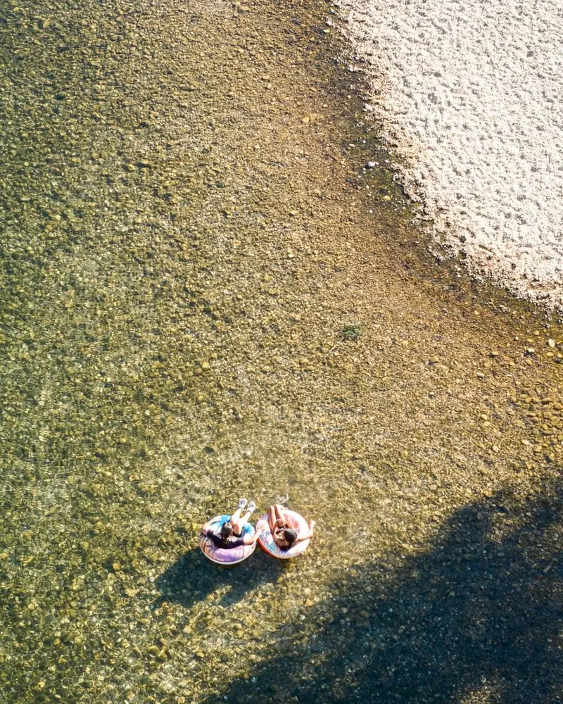 Two swimmers on inflatable rings floating over the clear, pebbly water of a river in the Gard, seen from above