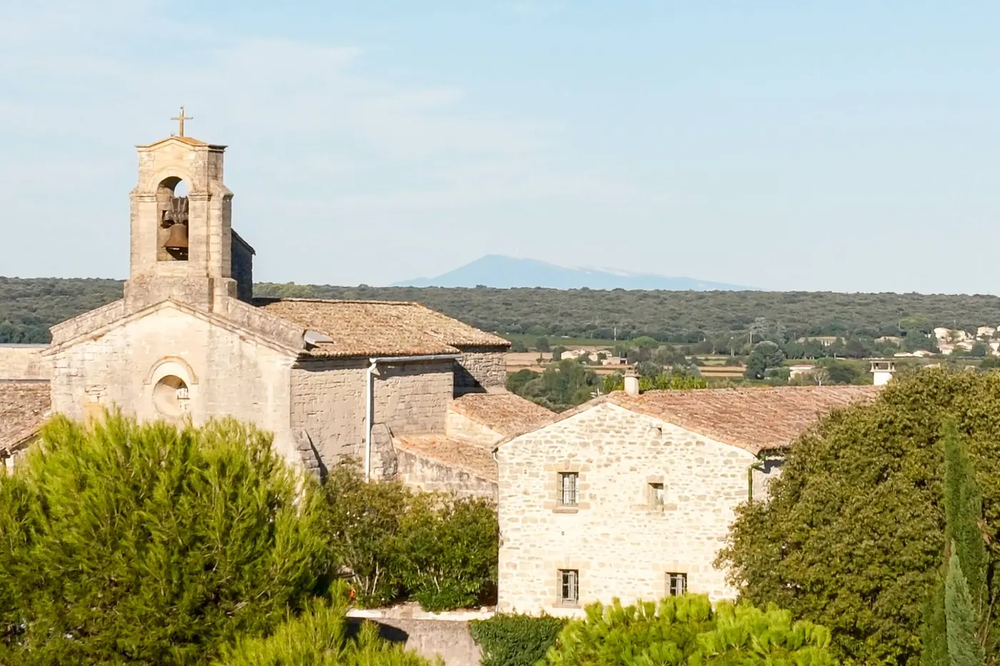 A village church with a stone bell tower among pines and cypresses, with hills and a distant peak on the horizon
