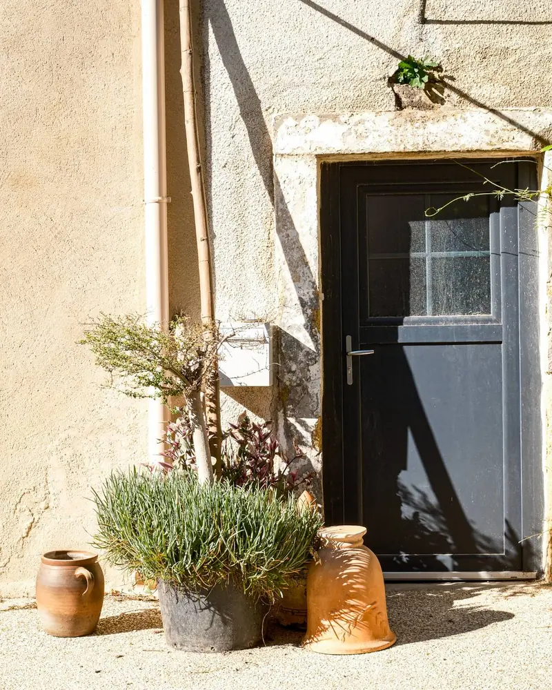 A dark door in a sunlit ochre wall, with terracotta pots and a pot of grasses by the step