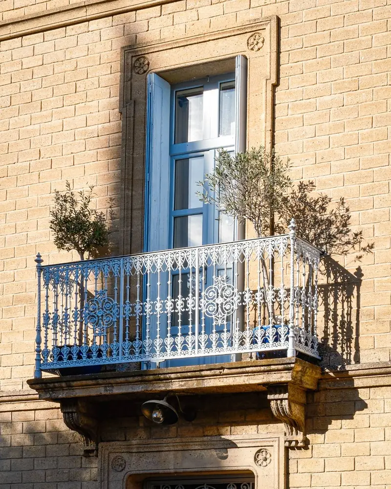 A wrought-iron balcony with pale blue shutters and two potted olive trees on a honey-coloured stone façade