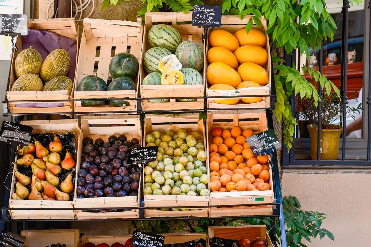 Wooden crates of melons, pears, plums and apricots under climbing greenery, with chalkboard labels reading Provence