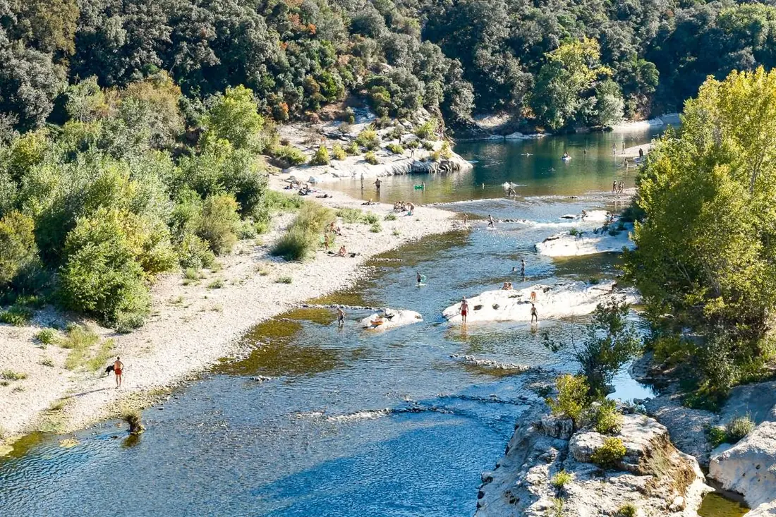 A river in the Gard with pale gravel banks and white rock slabs below wooded hills, with swimmers in the water