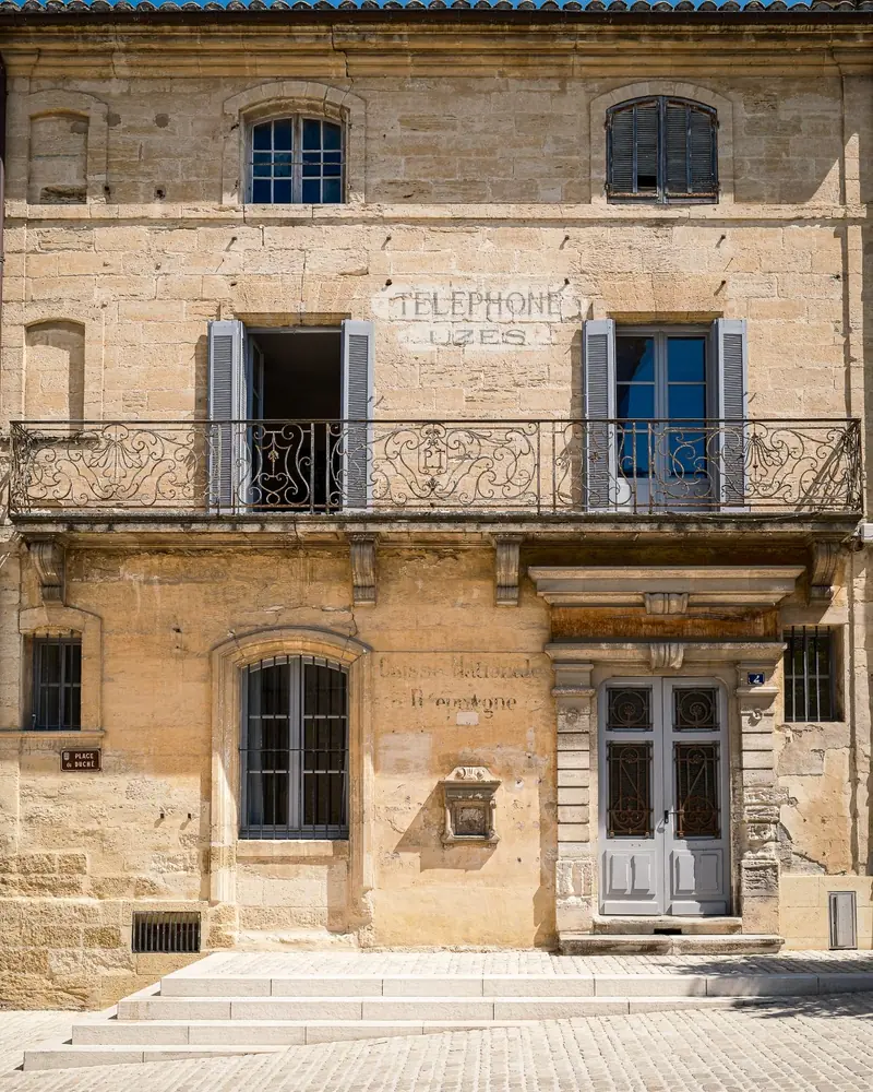 A honey-coloured stone façade on the Place du Duché in Uzès, with a wrought-iron balcony and an old painted telephone sign