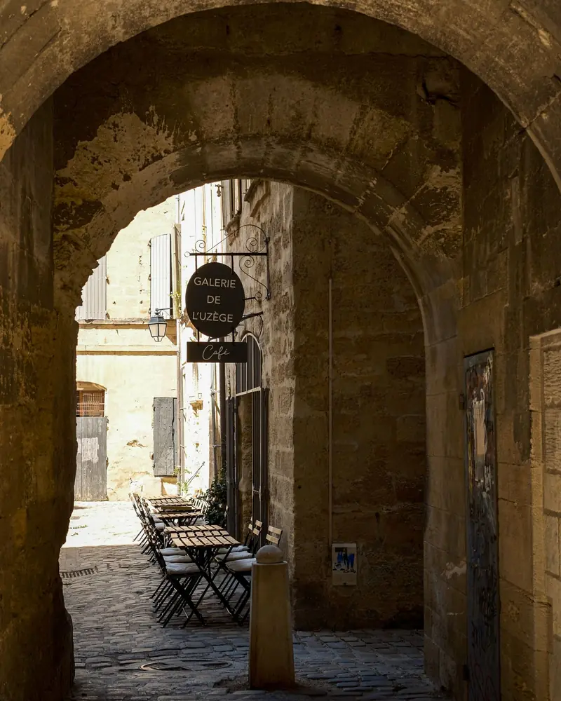 A vaulted stone passage in Uzès opening onto a sunlit courtyard with café tables